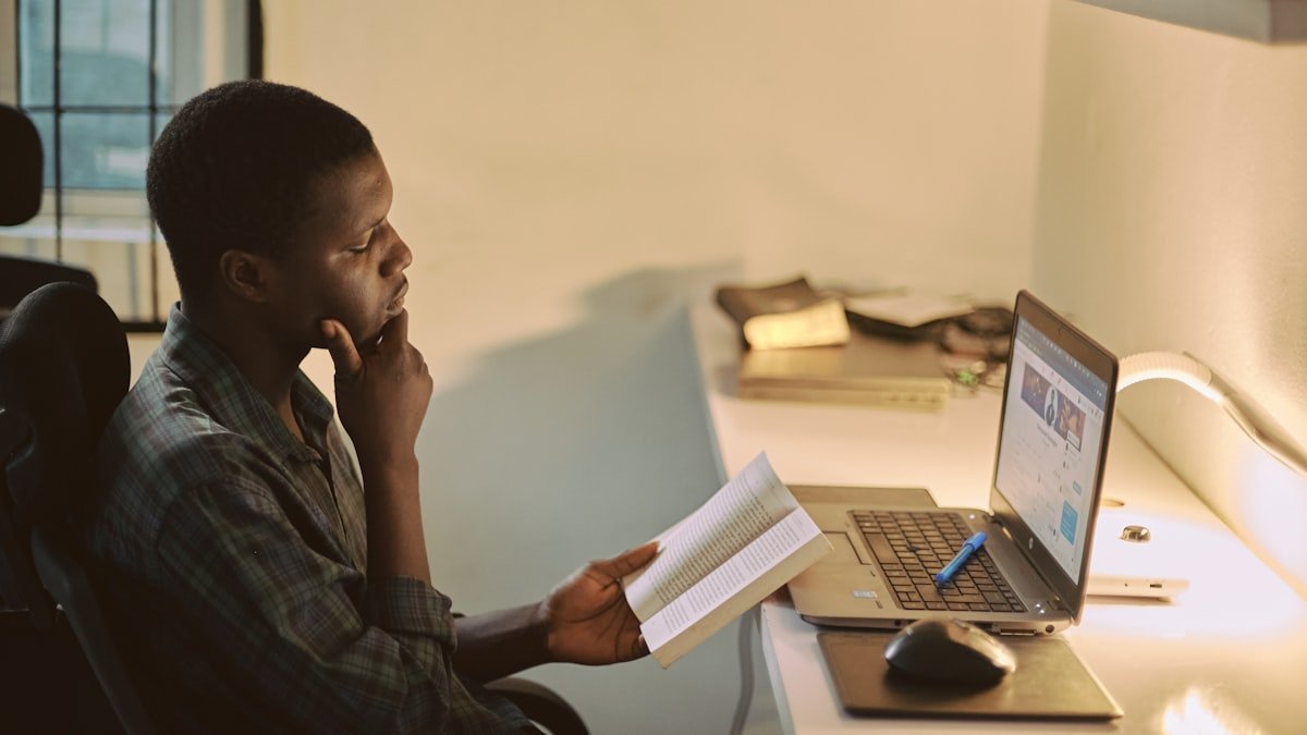A young Nigerian student sitting at an office desk, reading a printed document while checking his records