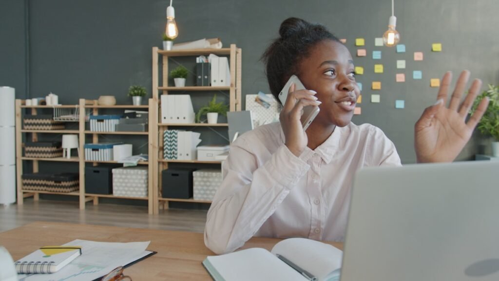 Young Nigerian graduate on the phone at her desk, box files behind her, chasing her NERD clearance record with her department
