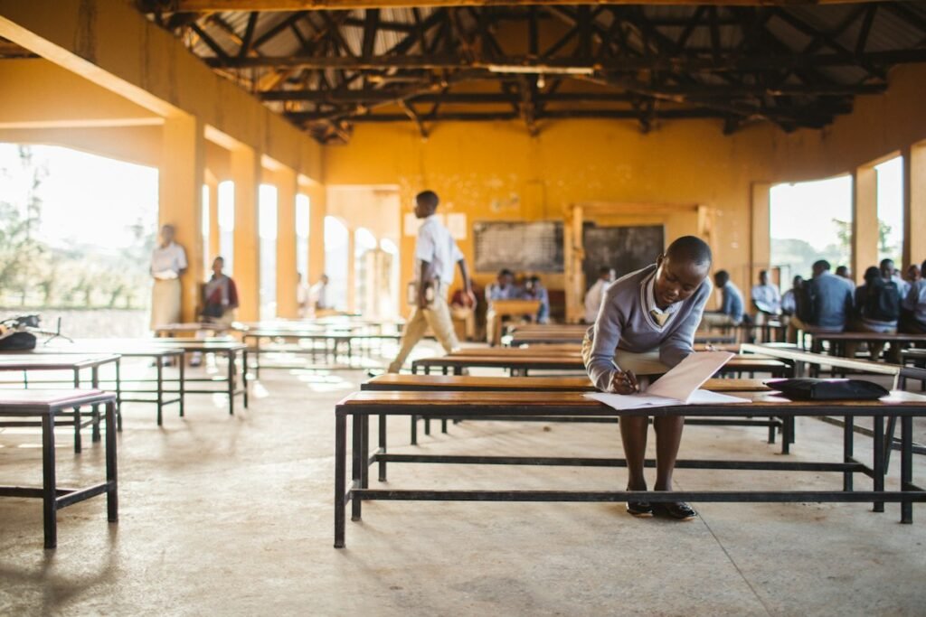 A secondary school student writing an exam paper at a bench in a school hall, the kind of setting NECO GCE 2026 candidates will sit in