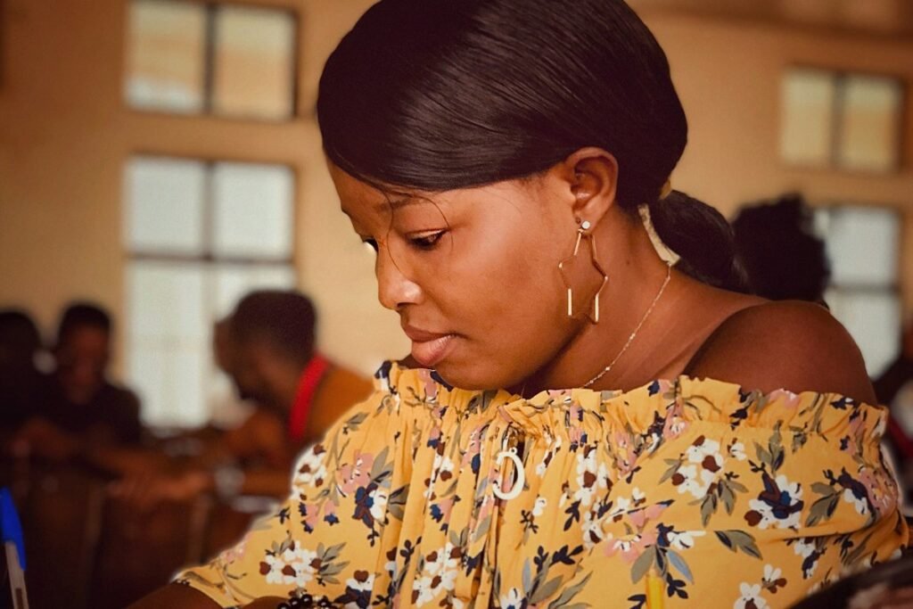 A young Nigerian woman writing at a desk in an exam hall, other candidates behind her